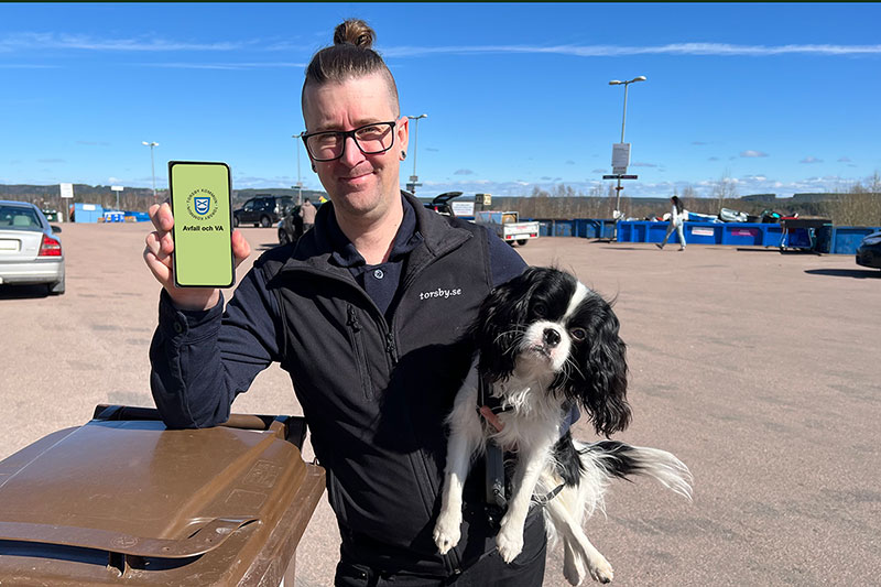 En man med en hund i famnen. Mannen står på en återvinningsstation. Han har en mobiltelefon i handen där man kan se en app.