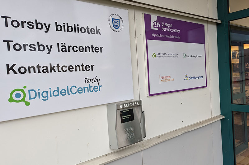 Utsidan på biblioteket med skylt Torsby bibliotek och under den är det en scanner, en silverfärgad låda.